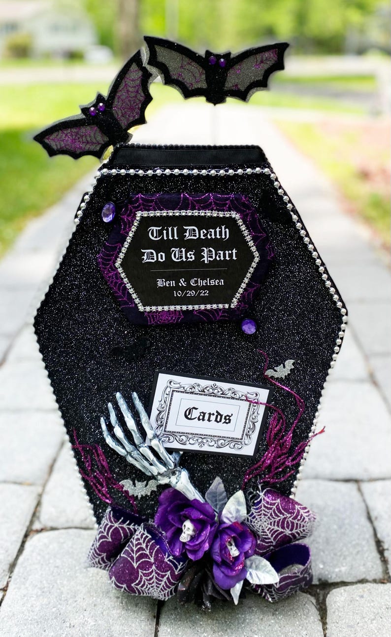 Puede incluir: Una caja de tarjetas negra en forma de ata&uacute;d con un acabado brillante y detalles morados. La caja tiene un letrero que dice "Till Death Do Us Part" con los nombres Ben & Chelsea y la fecha 10/29/22. La caja est&aacute; decorada con murci&eacute;lagos, telara&ntilde;as y flores.