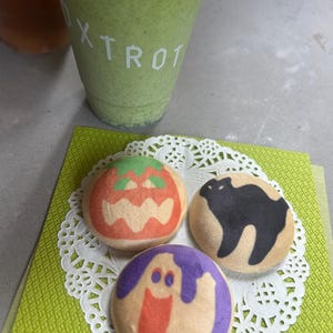 Pode incluir: Um smoothie verde em um copo de plástico transparente com a palavra "Xtrot" impressa. Três biscoitos redondos decorados com desenhos de Halloween estão em uma toalha de mesa branca e papel verde.