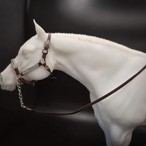 May include: A white horse figurine with a detailed bridle and reins. The bridle is brown leather with silver-colored decorative accents. A silver chain is attached to the bridle. The horse's head is turned to the left against a dark background.