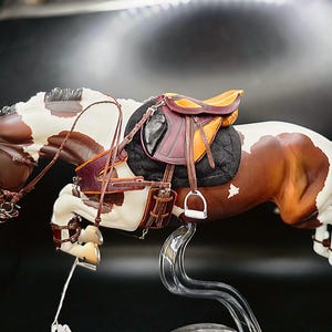 May include: A detailed figurine of a horse in mid-jump, showcasing a brown and white coat. The horse is equipped with a saddle, bridle, and reins. The saddle is a combination of black, brown, and yellow. The figurine is mounted on a clear stand.