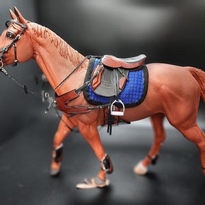 May include: A brown horse model with a black saddle and blue saddle pad. The horse is wearing a bridle and has black leg wraps.
