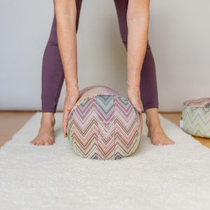 Könnte beinhalten: Ein zylindrisches Yoga-Bolster mit einem farbenfrohen Zickzackmuster in Rosa-, Grün- und Beigetönen. Das Bolster wird von einer Person in lila Leggings gehalten. Das Bolster liegt auf einem weißen Teppich.