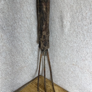 May include: A hand tool featuring a weathered, dark brown wooden handle and three thin, rusted metal prongs extending from the base. The handle shows a rough, aged texture with visible grain and vertical grooves near the top, while the metal tines are straight and pointed at the ends.
