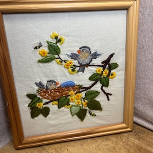 May include: A square framed embroidery piece on white fabric featuring two grey birds with orange beaks perched on brown branches with green leaves and yellow flowers. One bird sits in a brown nest with a blue egg, while the other perches nearby.