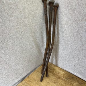 Puede incluir: Tres clavos metálicos largos y delgados con una gruesa capa de óxido marrón. Cada clavo presenta una cabeza plana y circular con un eje alargado ligeramente curvado. Las superficies metálicas muestran una oxidación significativa.