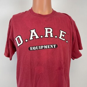 May include: A maroon short-sleeved t-shirt with the text "D.A.R.E. EQUIPMENT" in white and black lettering. The shirt is displayed on a mannequin and has a crew neck.