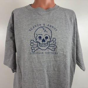 May include: Gray t-shirt with a navy blue skull and crossbones graphic. The graphic says "Search & Annoy" above the skull and "Dorothys Fortress" below.