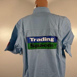 May include: Light blue short-sleeved shirt with the words "Trading Spaces" printed on the back in white, blue, and green. The shirt has a button-down collar and short sleeves. The shirt is displayed on a mannequin.