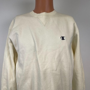 May include: A cream-colored long-sleeved sweatshirt with a small navy blue Champion logo on the left chest.