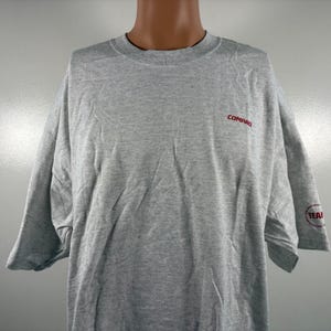 Peut inclure: Un t-shirt gris chiné à manches courtes. Le mot "COMPAQ" est imprimé en rouge sur le haut de la poitrine. La manche comporte un logo circulaire rouge avec le mot "TEAM" imprimé dessus. Le t-shirt est présenté sur un mannequin.