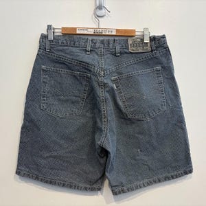 May include: Blue and white striped denim shorts with the Levi Strauss SilverTab label. The shorts have two back pockets with the Levi's logo and a button and zip closure. The shorts are hanging on a wooden hanger.