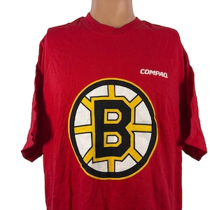 May include: Red t-shirt featuring the Boston Bruins logo in a black, yellow, and white design. The word "COMPAQ" is printed in white on the upper right. This vintage tee is a great addition to any hockey fan's wardrobe.