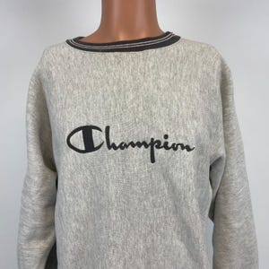 Pode incluir: Uma camisola cinzenta Champion com um logotipo bordado a preto na frente. A camisola tem um colarinho riscado preto e branco.
