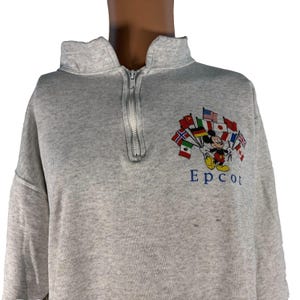 Puede incluir: Sudadera gris con cuello con cremallera. La sudadera presenta una imagen bordada de Mickey Mouse sosteniendo banderas de varios países, con la palabra "Epcot" en azul debajo.
