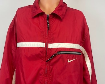 Puede incluir: Una chaqueta cortavientos roja con rayas blancas y un logotipo Nike Swoosh negro. La chaqueta tiene una cremallera y un cuello alto.