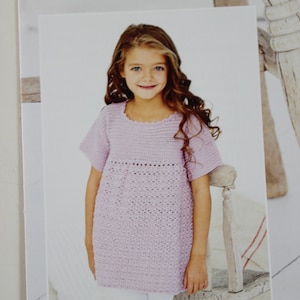May include: A young girl wearing a pale purple crocheted top with a scalloped neckline and short sleeves. She is standing in front of a white wooden chair.