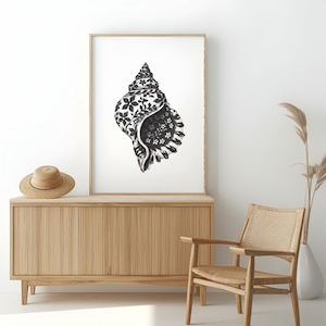 May include: A framed black and white seashell art print. The seashell is intricately detailed with floral patterns. The artwork is displayed above a wooden cabinet with a straw hat on top. A woven chair and vase with dried plants are also in the image.