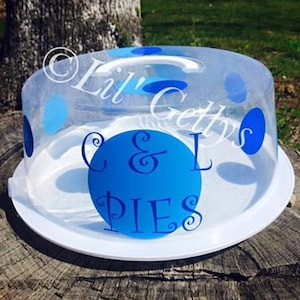 Puede incluir: Una cúpula para pastel de plástico transparente con un patrón de lunares azules y blancos. La cúpula tiene una manija blanca y una base blanca. El texto "C & L PIES" está escrito en azul en la cúpula.