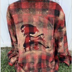 May include: Red and black plaid flannel shirt with a distressed design. The back features a silhouette of a figure in a Santa hat holding a gift. The shirt is displayed on a black mannequin head and set against a green grass background.