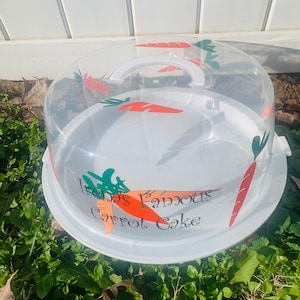 Könnte beinhalten: Ein weißer Kuchenbehälter mit einem durchsichtigen Deckel und einem Griff. Der Behälter hat ein Karotten-Design und den Text "Irene's Famous Carrot Cake".