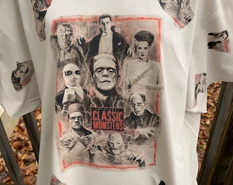Pode incluir: Uma camiseta branca com uma estampa gráfica de personagens clássicos dos monstros universais, incluindo Frankenstein, Drácula, a Múmia e o Lobisomem. O gráfico é cercado por uma borda vermelha e inclui o texto "Universal Classic Monsters".