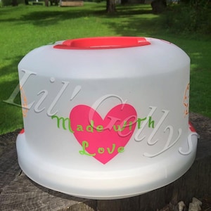 Puede incluir: Un porta tartas blanco con una tapa rosa y un corazón rosa con el texto "hecho con amor" en letras verdes.