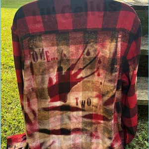 May include: Red and black plaid flannel shirt with a distressed graphic on the back. The graphic features a silhouette of a clawed hand with the words "ONE... TWO..." in a distressed font. The shirt is button-down.