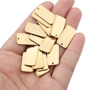 Könnte beinhalten: Eine Sammlung kleiner, rechteckiger, goldfarbener Metallanhänger mit abgerundeten Ecken und einem einzelnen Loch oben. Die Anhänger werden in einer Hand vor weißem Hintergrund gehalten. Diese Anhänger eignen sich für die Schmuckherstellung oder zum Basteln.