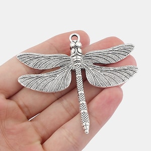 Op de afbeelding: Zilverkleurige libellenbedel met gedetailleerde vleugels en een gestructureerd lichaam. De bedel heeft een lus aan de bovenkant om aan sieraden of andere knutselwerken te bevestigen.