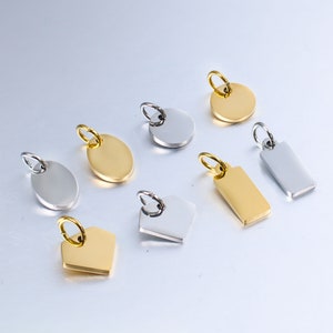 May include: A set of eight blank metal charms in silver and gold. The charms are in various shapes, including circles, ovals, rectangles, and a diamond shape. The charms have a hole at the top for attaching to a necklace or bracelet.