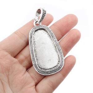 May include: Silver pendant with a large oval-shaped bezel. The bezel has a decorative border and is ready to be filled with a stone or other material.