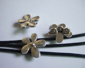 May include: Three silver-colored flower-shaped clasps. The clasps are made of metal and have a detailed design. The clasps are on a black leather cord. The background is white.
