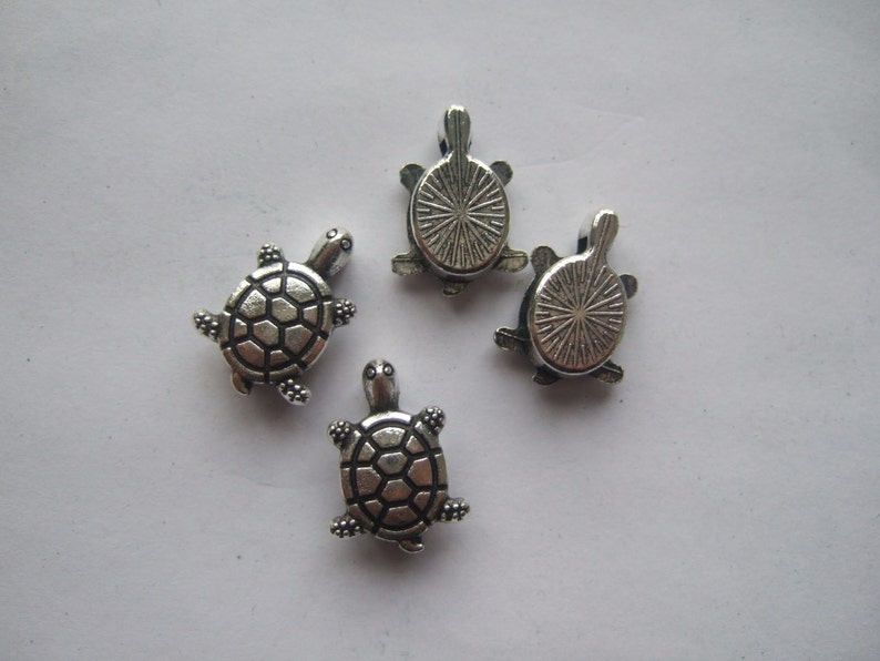 May include: Four silver turtle charms with detailed shell designs. The turtles are about 1 inch in size and have a loop at the top for attaching to jewelry or other crafts.