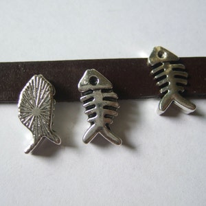 Puede incluir: Tres abalorios de metal plateado en forma de esqueletos de pescado. Los abalorios están en una correa de cuero marrón.
