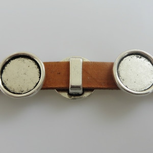 Peut inclure: Deux sertis ronds en métal argenté pour la fabrication de bijoux, fixés à une lanière en cuir marron. Les sertis sont conçus pour recevoir un cabochon rond.