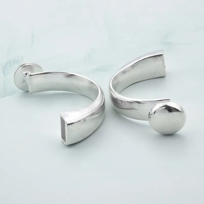 May include: A pair of silver hoop earrings with a rounded, smooth finish. The earrings have a unique design with a flat, rectangular section on one side and a rounded ball on the other.