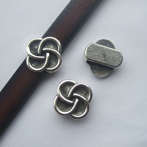 May include: Three silver metal Celtic knot charms, two are four-leaf clover shaped and one is a rectangular shape with a textured design.