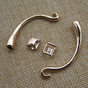 Puede incluir: Dos asas de bolso de metal dorado con un rizo decorativo en el extremo. Las asas son curvadas y tienen un acabado liso. También hay dos pequeñas piezas de metal dorado, una con forma de estrella y la otra con forma de cuadrado.