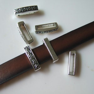 May include: Five silver metal belt buckle closures with a geometric pattern. The closures are rectangular and have a small opening in the center. The closures are on a brown leather strap.