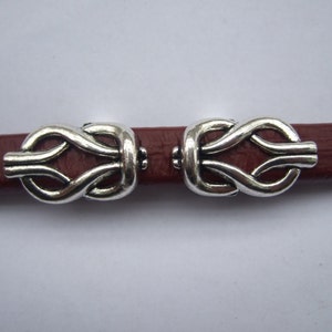 Pode incluir: Dois fechos de metal prateado com design de nó, mostrados em uma alça de couro marrom.
