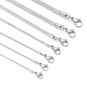 Puede incluir: Una colección de collares de cadena de serpiente plateados de varias longitudes. Cada collar presenta un cierre de mosquetón. Las cadenas se muestran sobre un fondo blanco, resaltando su brillo metálico y su intrincado diseño. Medidas en centímetros.