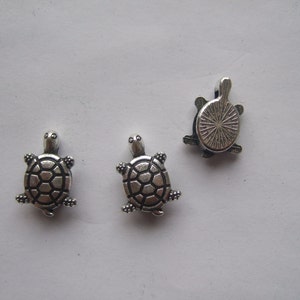 May include: Three silver turtle charms. Two of the turtles are facing forward with their legs and heads visible. The third turtle is facing backward with its shell visible.