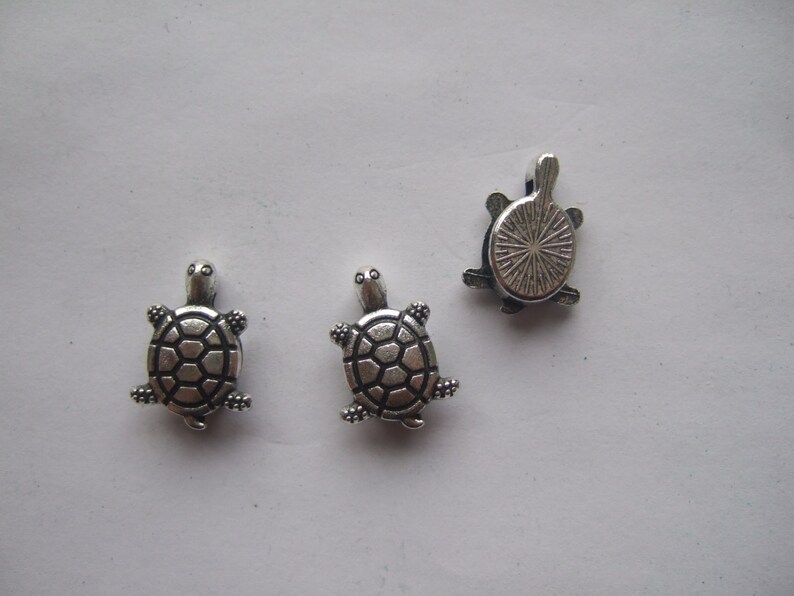 May include: Three silver turtle charms. Two of the turtles are facing forward with their legs and heads visible. The third turtle is facing backward with its shell visible.