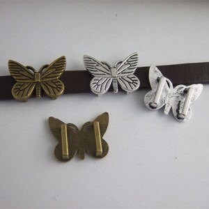 Op de afbeelding: Vier metalen vlinderversieringen in brons- en zilverkleuren. De vlinders zijn bevestigd aan een bruine leren riem. De vlinders hebben gedetailleerde vleugelpatronen en zijn ontworpen voor decoratieve doeleinden.