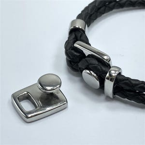 Puede incluir: Un brazalete de cuero negro con un cierre plateado y un cierre de palanca plateado. El brazalete está trenzado y tiene un acabado liso y pulido.