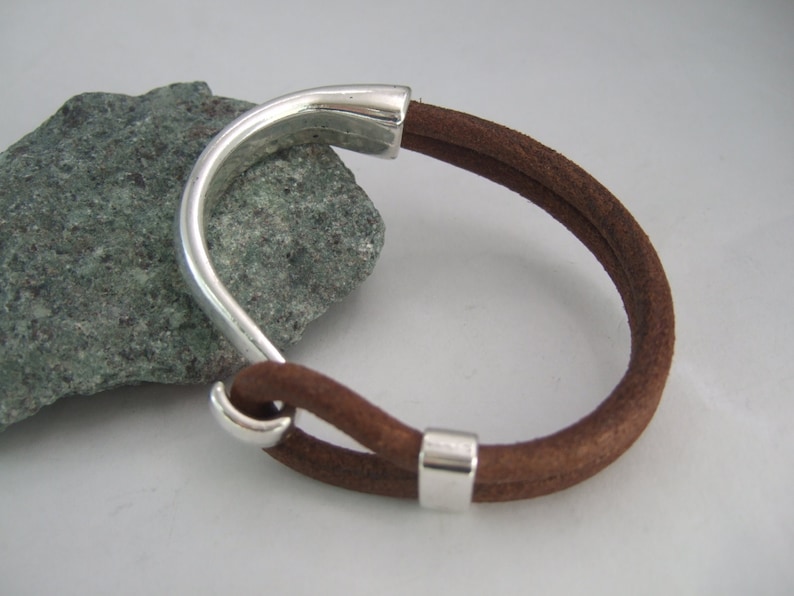 May include: A brown leather bracelet with a silver metal clasp. The bracelet is made of two strands of leather that are woven together.
