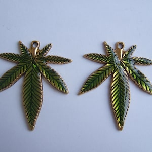 May include: Two gold-colored metal charms shaped like marijuana leaves. The leaves are green with gold veins and have a shiny finish.