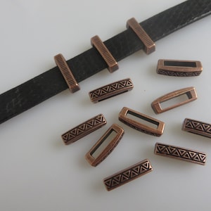 Peut inclure: Une lanière en cuir noir texturé avec trois passants métalliques rectangulaires couleur cuivre à bords striés, accompagnée de huit passants métalliques couleur cuivre supplémentaires présentant un motif triangulaire répété sur la face avant.