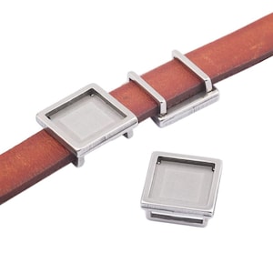 Puede incluir: Dos marcos cuadrados de metal plateados con un inserto de vidrio transparente. Los marcos están diseñados para ser utilizados como un amuleto en una pulsera de cuero.