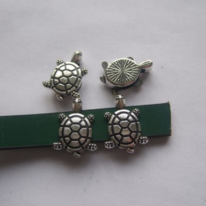 May include: Four silver turtle charms with detailed shell designs. Two turtles are attached to a green leather strap.